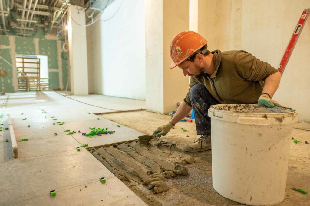 A construction worker lays tiles in an indoor renovation project, wearing safety gear.